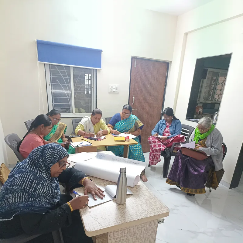 Eight women seated around tables in a meeting room, writing notes, beside a chart paper headed Election Campaign.