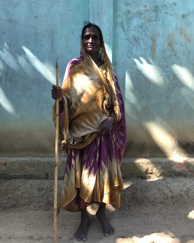 A Dalit elder in a patterned sari stands in the sun against a pale blue wall, holding a bamboo walking stick and looking at the camera.