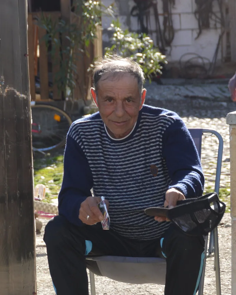 A Roma elder seated outdoors in a sunlit yard, looking directly at the camera.