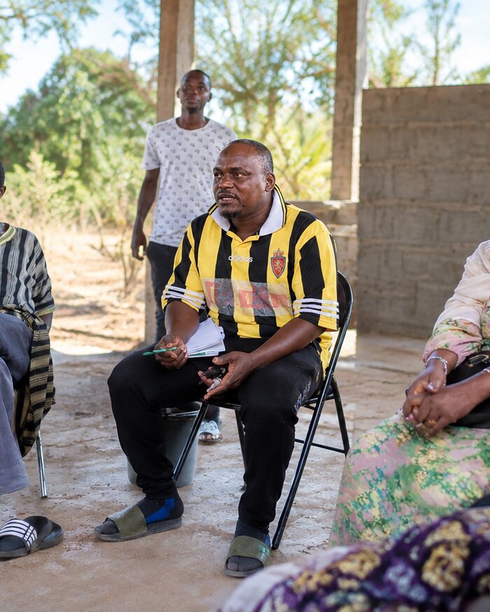 Ali Camarra speaking at a community meeting in The Gambia, seated in a yellow and black striped shirt.