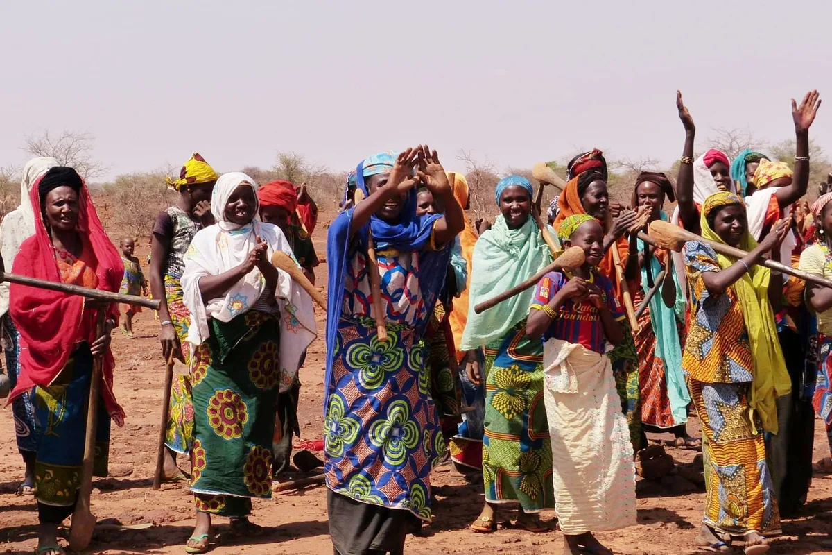 A crowd of Haratine women in bright headscarves and patterned wraps, some carrying long wooden tools, clapping and waving on open red earth.