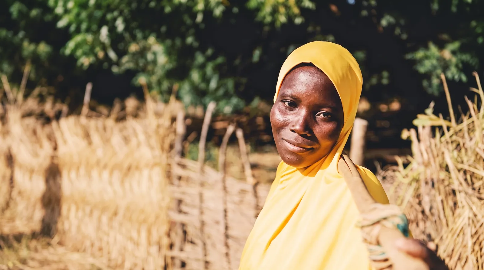 A young woman in a yellow headscarf smiles at the camera, a wooden tool over her shoulder, beside a woven straw fence.