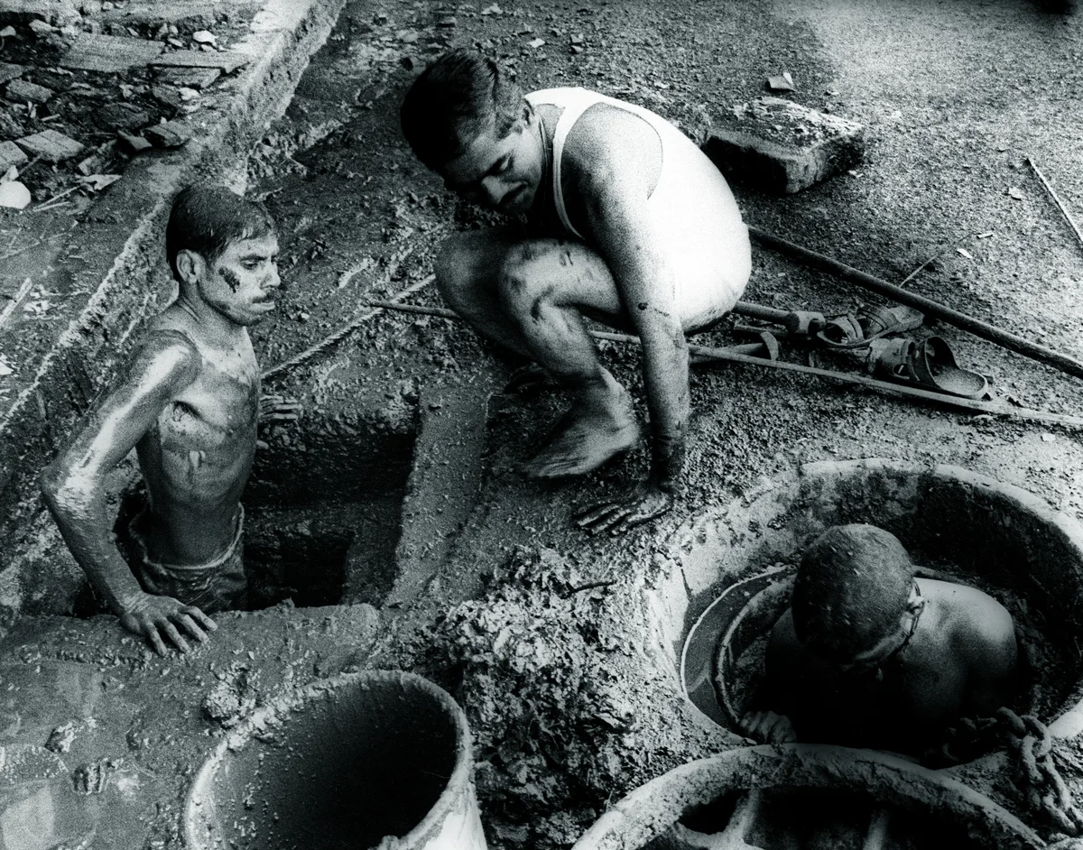 Two men stand chest-deep in open sewer pits, covered in sludge, while a third crouches at the edge beside them.