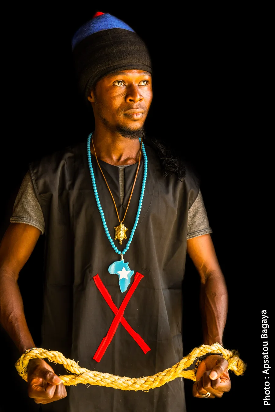 A young man in a dark tunic, with a red cross marked on his chest and an Africa pendant, holds a length of rope stretched between his hands.