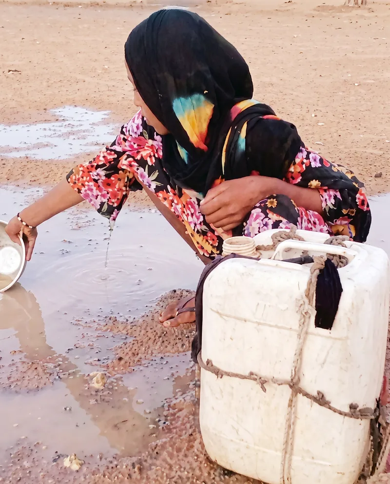 A woman crouches to fill a jerrycan from a shallow pool of standing water.