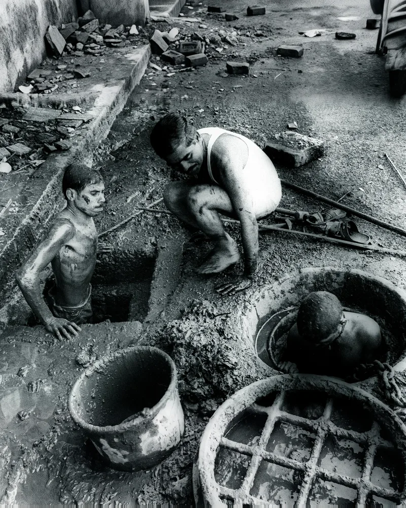 Two men stand chest-deep in open sewer pits, covered in sludge, while a third crouches at the edge beside them.
