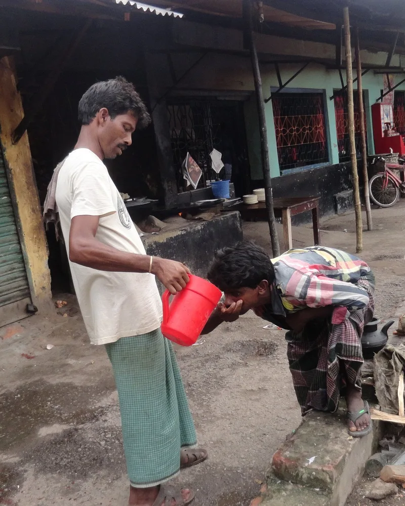 A man pours water from a jug directly into another man&rsquo;s cupped hands in a street in Bangladesh.