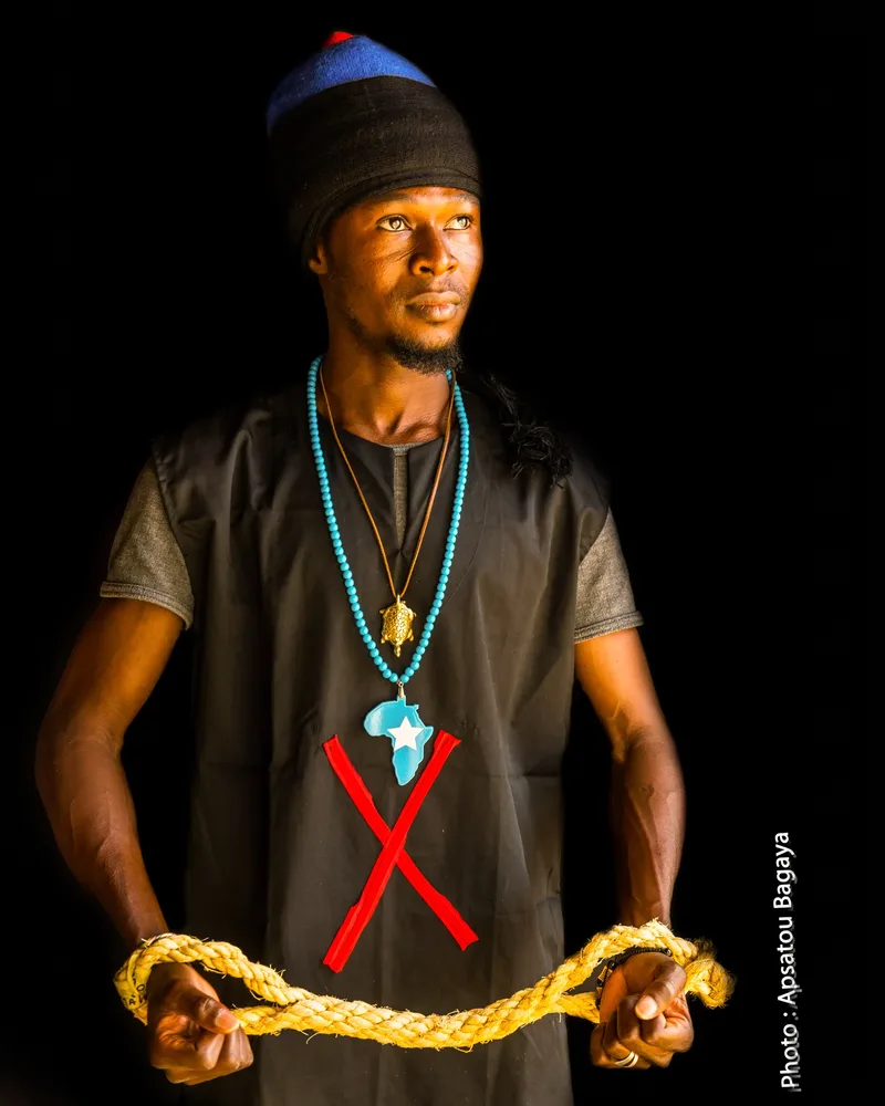 A young man in a dark tunic, with a red cross marked on his chest and an Africa pendant, holds a length of rope stretched between his hands.