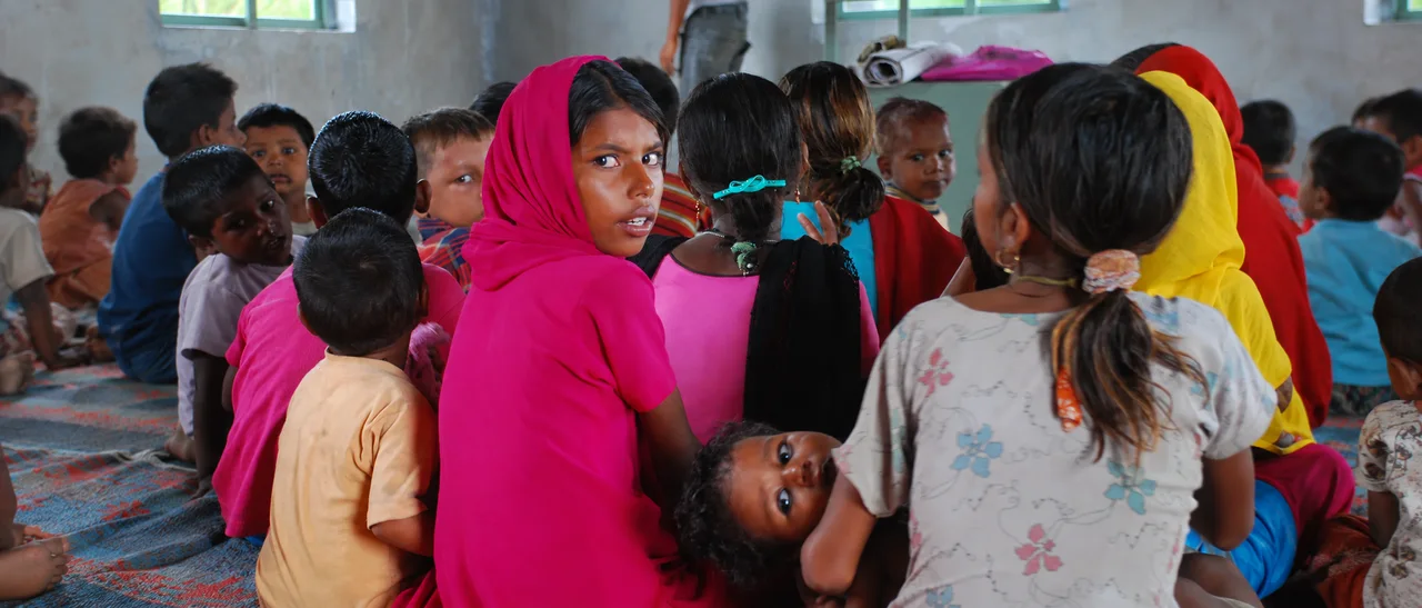 A girl in a pink headscarf turns to look back over her shoulder, sitting among children on the floor of a classroom.