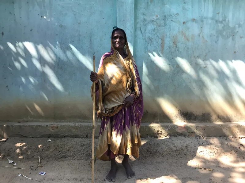 An elderly woman in a patterned sari stands with a bamboo stick against a pale blue wall.
