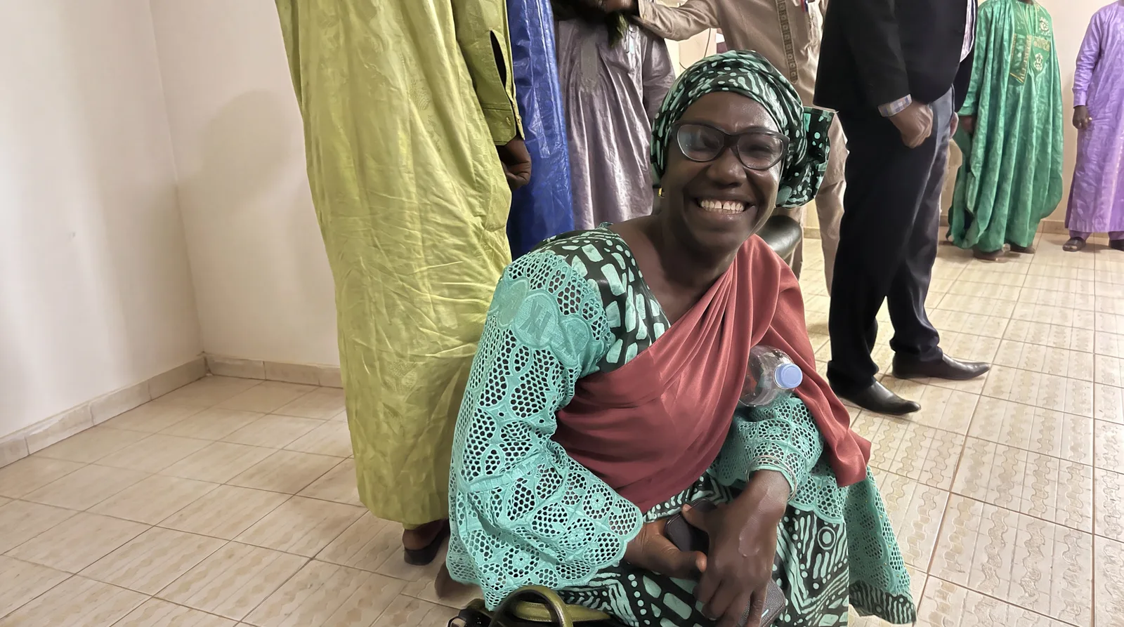 A smiling woman in glasses and a green patterned headwrap, wearing a turquoise lace dress with a deep red sash, seated among colleagues.