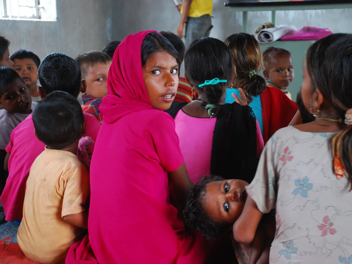A girl in a pink headscarf looks back over her shoulder, a baby resting against her, among children sitting together in a classroom.
