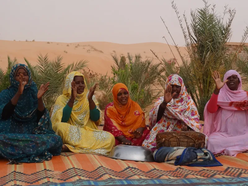 Five women in bright headscarves sing and clap together on a woven mat in the desert.