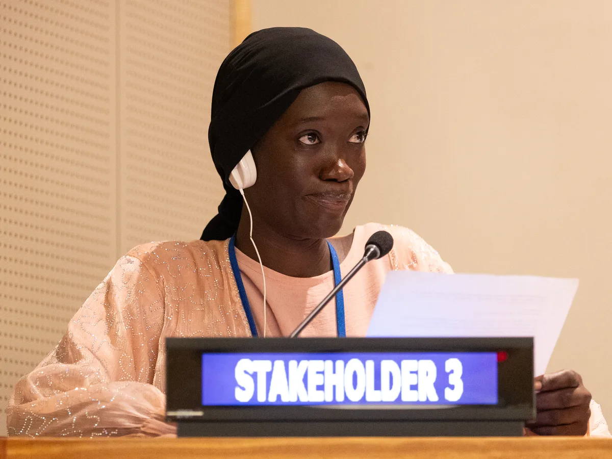 A young woman in a black headscarf and interpreter earpiece speaks at a UN stakeholder desk, notes in hand.