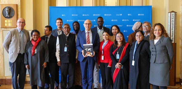 A group of eighteen community members and colleagues standing in a row with Volker T&uuml;rk, who holds a report, in front of a blue UN Human Rights backdrop in Geneva.
